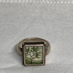 May include: A silver ring with a square-shaped setting featuring a green tree with a red house in the background. The ring is on a white fabric background.