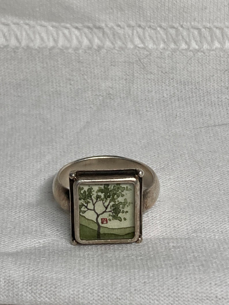 May include: A silver ring with a square-shaped setting featuring a green tree with a red house in the background. The ring is on a white fabric background.