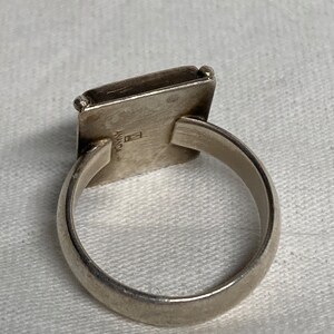 May include: A silver ring with a square-shaped top. The top of the ring has a small square with the word "ANANDA" inscribed on it.