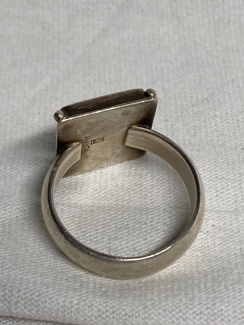 May include: A silver ring with a square-shaped top. The top of the ring has a small square with the word "ANANDA" inscribed on it.