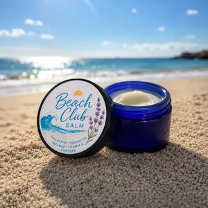 May include: A cobalt blue glass jar containing off-white balm with a black lid featuring a label that reads Beach Club Balm, with graphics of a sun, a wave, and lavender sprigs, listing shea butter, coconut oil, beeswax, vitamin E, and lavender.