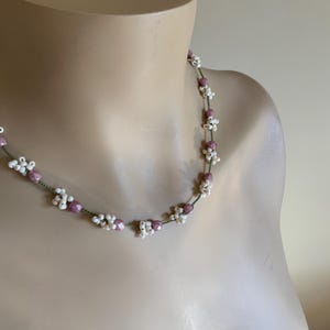 May include: A delicate necklace featuring a string of small, white, flower-shaped beads interspersed with larger, round, pink beads. The beads are strung on a thin, green cord, creating a floral pattern. The necklace is displayed on a mannequin.
