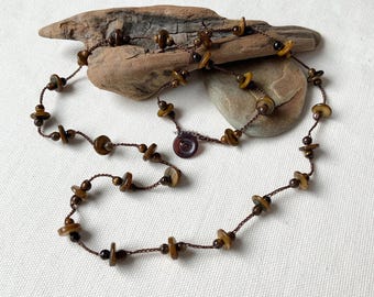 May include: A brown beaded necklace with a button closure. The necklace features small, round, and disc-shaped beads in shades of brown and tan, strung on a dark brown cord. The necklace is displayed on a piece of driftwood.