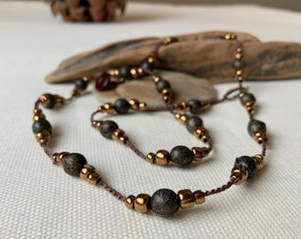 May include: A necklace and bracelet set featuring dark green and gold-colored beads strung on a brown cord. The beads have a speckled appearance. The necklace and bracelet are displayed on a white surface.