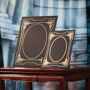 Peut inclure: Deux cadres noirs avec une bordure décorative dorée et des ouvertures ovales. Les cadres sont exposés sur une table en bois foncé. Les cadres ont un motif géométrique dans les coins et sur les bords.
