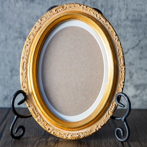 May include: Oval gold picture frame with decorative detailing, supported by a black metal stand. The frame has a white inner border and a brown backing. The frame is displayed on a dark wood surface.