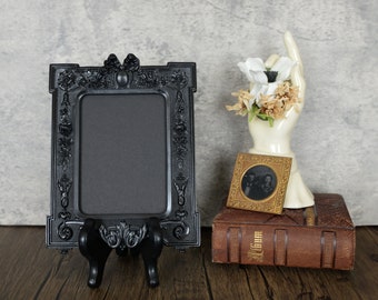 May include: Black ornate picture frame with a black easel stand. The frame is empty and has a decorative design. A small gold picture frame with a black and white photo is sitting on top of a brown leather-bound book.