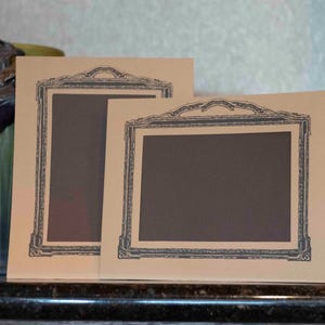 Pode incluir: Dois cartões retangulares de papel bege, cada um com uma ilustração impressa em cinza escuro de uma moldura decorativa e ornamentada. Os cartões estão posicionados na vertical, um orientado verticalmente e o outro horizontalmente.