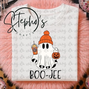 May include: White t-shirt featuring a cartoon ghost wearing an orange beanie and holding a pumpkin bucket and a coffee drink. The text "BOO-JEE" is printed below the ghost. The shirt has the logo "Stepho's Crafts" at the top.