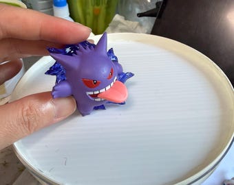 May include: A small purple Gengar figurine with pointed ears, red eyes, and a long pink tongue sticking out. The character is surrounded by translucent dark blue energy effects and is held against a white circular surface.