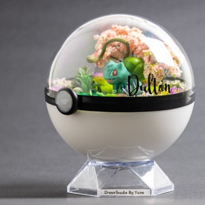 Peut inclure: Un terrarium blanc et noir en forme de Poké Ball, avec un dôme transparent. À l'intérieur, une figurine verte de Bulbasaur est entourée de plantes miniatures et de fleurs roses. Le nom "Dalton" est écrit sur le dôme.