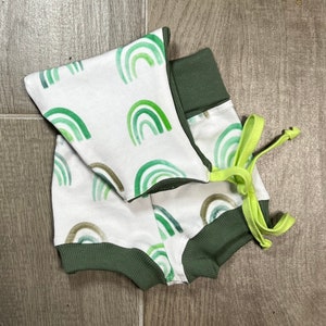Puede incluir: Un pelele blanco para bebé con estampado de arcoíris acuarela verde y ribete verde. El pelele tiene un gorro a juego verde y blanco con una corbata verde.