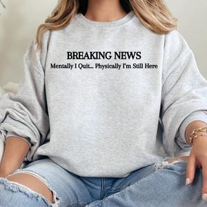 Puede incluir: Una sudadera gris claro con el texto negro "BREAKING NEWS Mentally I Quit... Physically I'm Still Here". La sudadera se combina con unos vaqueros azules.