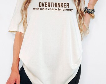 May include: Cream-colored t-shirt with the text "OVERTHINKER with main character energy" in brown. The shirt is a relaxed fit. The image also shows black shorts.