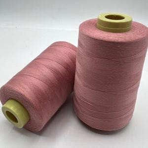 May include: Two spools of pink thread. The thread is tightly wound around a yellow core. The spools are cylindrical and appear to be made of a smooth, shiny material. The thread is a muted pink colour.