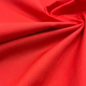 May include: A vibrant red fabric with a smooth, matte texture. The material is draped in soft, flowing folds that create subtle shadows and highlights across the surface, showcasing the consistent, solid colour and fine weave of the textile.