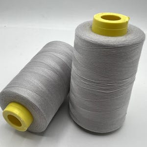 May include: Two spools of grey thread with yellow cores are shown against a white backdrop. The thread is tightly wound, and the spools are cylindrical. The image is well-lit, highlighting the texture of the thread.