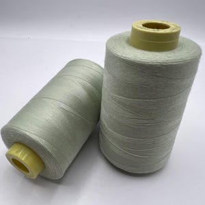 May include: Two spools of pale green thread are shown against a white backdrop. The thread is tightly wound around a cylindrical core, with a yellow cap on each spool. The thread has a slight sheen.
