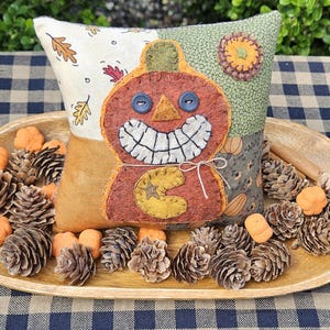 May include: A square fabric pillow featuring a patchwork design with cream, green, and mustard yellow sections. A central felt pumpkin applique has a wide stitched grin, blue button eyes, and a yellow crescent moon shape with a star cutout.