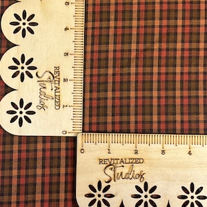 May include: Two wooden rulers with floral cutouts and the text "REVITALIZED Studios" are positioned at a right angle against a brown, orange, and black plaid fabric. The rulers have inch markings.