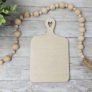 May include: A light-colored wooden cutting board with a heart-shaped cutout in the handle. A decorative wooden bead garland and greenery are in the background. The cutting board is approximately 10 inches long.