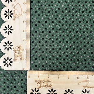 May include: Close-up of a green fabric with a small floral pattern. Two wooden rulers with scalloped edges and laser-cut flower designs are positioned at a right angle. The rulers have the text "REVITALIZED Studios" and "From" on them.