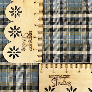 May include: Two wooden rulers with a floral design and the text "Revitalized Studios" are placed over a plaid fabric. The fabric features a pattern of intersecting lines in shades of blue, green, black, and white. The rulers measure in inches.