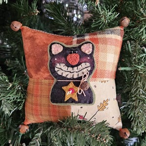 May include: A square fabric ornament featuring a patchwork design in orange, brown, and cream tones. A black felt cat appliqué with a wide stitched grin, a red nose, and a yellow star sits in the center. Small metal bells are attached to each corner.