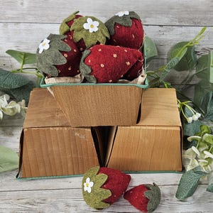 Könnte beinhalten: Ein hölzerner Beerenkorb gefüllt mit handgefertigten roten Filz-Erdbeeren mit grünen Blättern und weißen Blumenakzenten. Zwei zusätzliche Erdbeeren sind vor dem Korb platziert. Die Erdbeeren liegen auf einer Holzoberfläche.