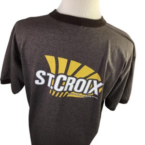 May include: A dark gray t-shirt with a brown collar and sleeve trim. The shirt features the white text "ST. CROIX" with a yellow sunburst design. Below the text is the phrase "HANDCRAFTED FISHING RODS".