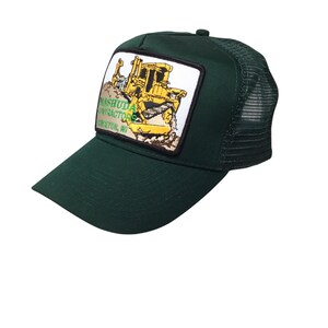 May include: A dark green trucker hat with a mesh back and a curved peak. The front has a rectangular patch with a yellow bulldozer graphic and the text "Mashuda Contractors, Princeton, WI."
