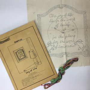 Puede incluir: Un kit de bordado beige con un diseño de sampler que dice "The joy of life lies in the work of today and in the plans for the work of tomorrow." El kit incluye una pieza de tela beige, hilo de bordar en varios colores y una hoja de instrucciones impresa.