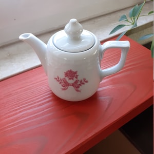 May include: A white ceramic teapot with a lid and handle, featuring a pink floral design with a bow. The teapot is placed on a red wooden surface, with a window and greenery in the background.
