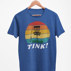 May include: A blue t-shirt with a retro sunset graphic featuring a disc golf basket and the text "TINK!"