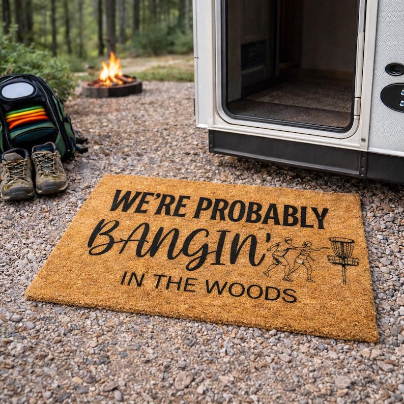 May include: A rectangular coir doormat with the text "WE'RE PROBABLY BANGIN' IN THE WOODS" in black script. The mat features a graphic of two people playing disc golf and a disc golf basket. The mat is placed outside a camper.