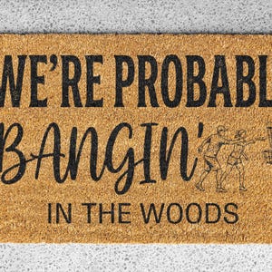 May include: Rectangular doormat with the text "WE'RE PROBABLY BANGIN' IN THE WOODS" in black. The mat features a line drawing of two people playing disc golf and a disc golf basket. The mat is made of a natural fiber.