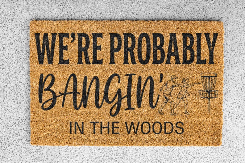 May include: Rectangular doormat with the text "WE'RE PROBABLY BANGIN' IN THE WOODS" in black. The mat features a line drawing of two people playing disc golf and a disc golf basket. The mat is made of a natural fiber.