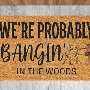 May include: A rectangular doormat made of natural coir fiber. The mat features the text "WE'RE PROBABLY BANGIN' IN THE WOODS" in black, along with a graphic of people playing disc golf.