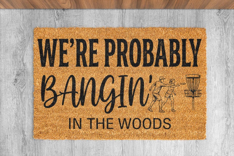 May include: A rectangular doormat made of natural coir fiber. The mat features the text "WE'RE PROBABLY BANGIN' IN THE WOODS" in black, along with a graphic of people playing disc golf.