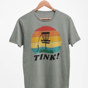 May include: A green t-shirt with a retro sunset graphic featuring a disc golf basket and the text "TINK!"