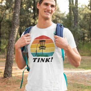 May include: A white t-shirt with a retro-style graphic of a disc golf basket and the text "TINK!" printed on the front.