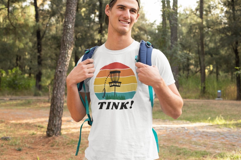 May include: A white t-shirt with a retro-style graphic of a disc golf basket and the text "TINK!" printed on the front.