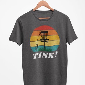 May include: A dark heather grey t-shirt with a retro-style sunset graphic featuring a disc golf basket and the text "TINK!"