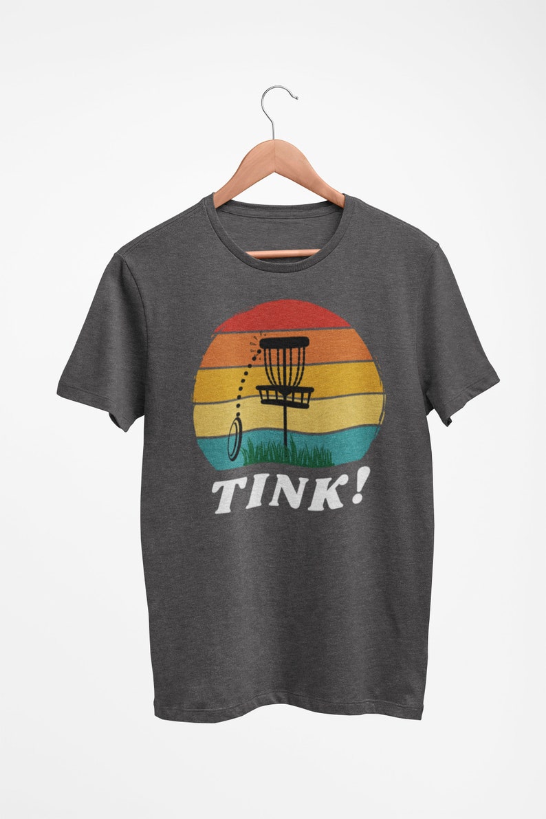 May include: A dark heather grey t-shirt with a retro-style sunset graphic featuring a disc golf basket and the text "TINK!"