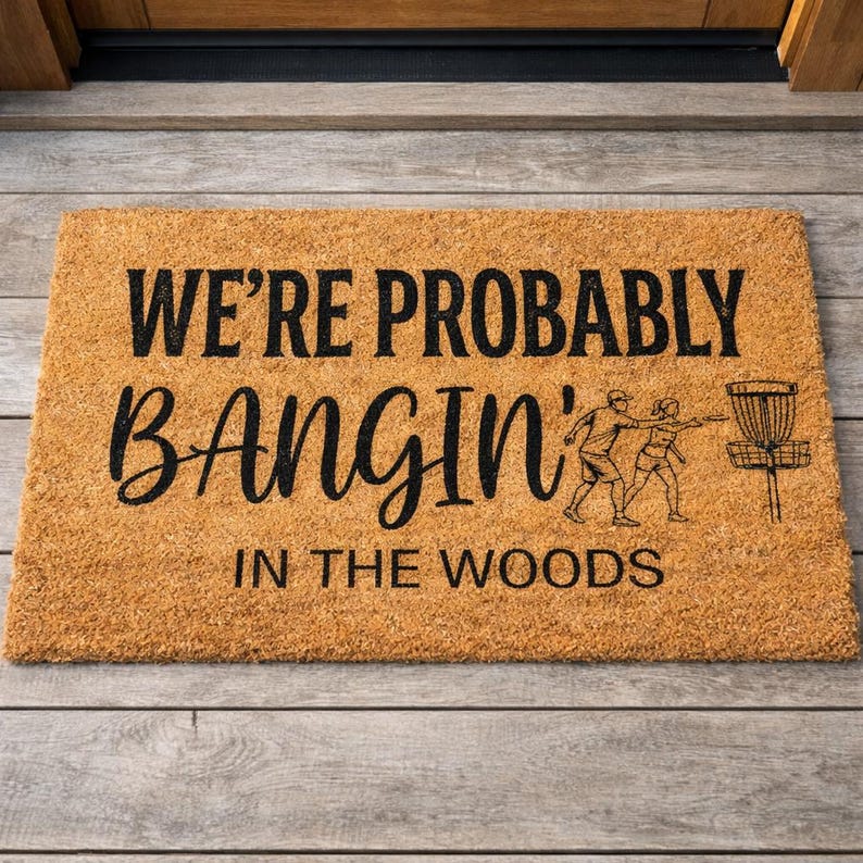 May include: A rectangular doormat made of natural coir fiber. The mat features the text "WE'RE PROBABLY BANGIN' IN THE WOODS" in black, along with a graphic of two people playing disc golf and a disc golf basket.