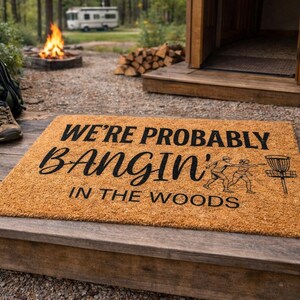 May include: A rectangular coir doormat with the text "WE'RE PROBABLY BANGIN' IN THE WOODS" in black. The mat features a graphic of two people playing disc golf. Hiking boots, a disc golf bag, and a campfire are in the background.
