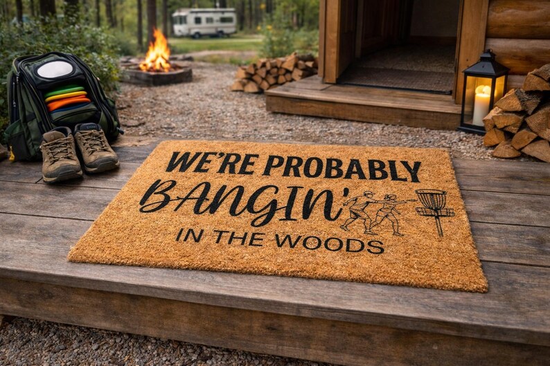 May include: A rectangular coir doormat with the text "WE'RE PROBABLY BANGIN' IN THE WOODS" in black. The mat features a graphic of two people playing disc golf. Hiking boots, a disc golf bag, and a campfire are in the background.