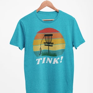 May include: A turquoise blue t-shirt with a retro sunset graphic featuring a disc golf basket and the text "TINK!"