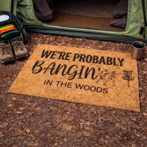 May include: A rectangular coir doormat with the text "WE'RE PROBABLY BANGIN' IN THE WOODS" in black. A graphic of two people playing disc golf is on the right. Hiking boots, a backpack, and a tent are in the background.