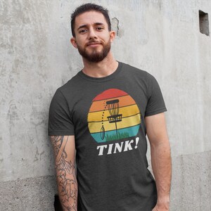 May include: A dark grey t-shirt with a retro-style graphic of a disc golf basket in front of a sunset. The text "TINK!" is printed below the graphic.